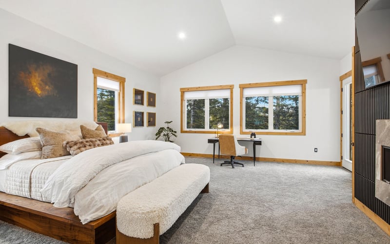 A bedroom with a king bed, cream bedding, a textured bench, dark wood accents, and a desk under windows in a bright room.