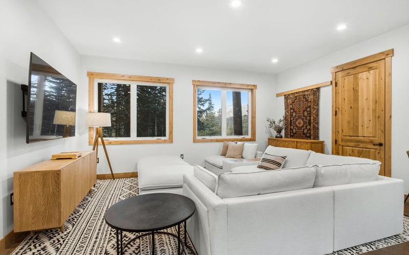 A bright living room with a white sectional sofa, a patterned rug, a wooden media console, and two windows with wood trim.