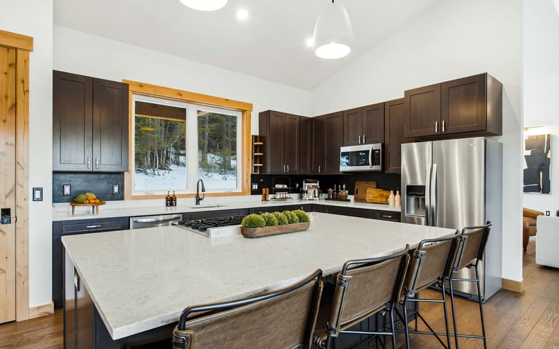 A bright, modern kitchen featuring dark wood cabinets, a large white island with seating, and a window with snowy views.