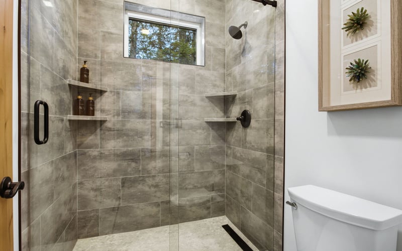 A modern walk-in shower with grey stone-look tile, glass door, built-in shelves, and a toilet in a white bathroom.