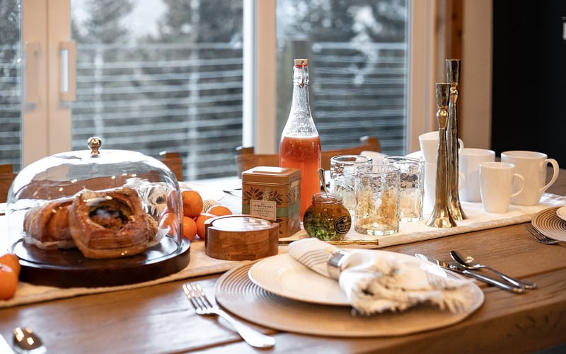 A breakfast table setting with pastries under a glass dome, orange juice, tea, and place settings near a bright window.