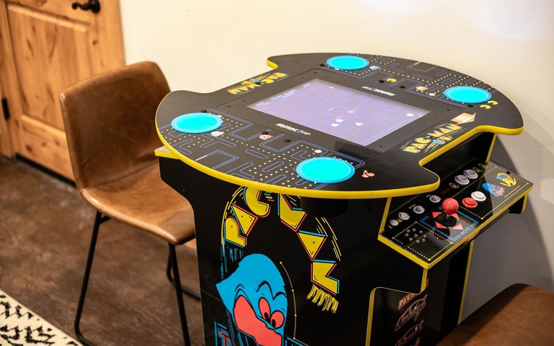 A Pac-Man arcade cocktail table with blue illuminated buttons, situated in a room next to a brown chair.