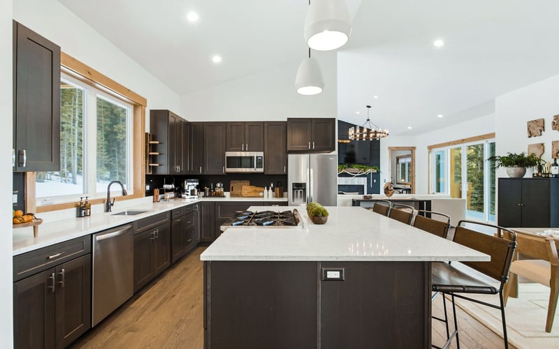 Modern kitchen with dark wood cabinets, a white island, stainless steel appliances, and large windows looking outside.