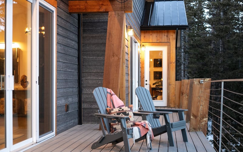 Two Adirondack chairs with blankets sit on a wooden deck outside a modern house with wood siding, overlooking a forest.