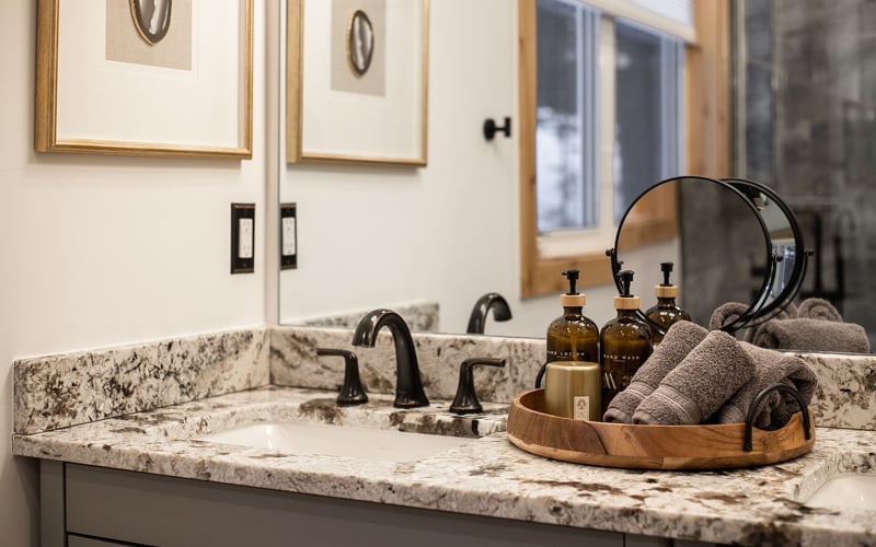 A granite bathroom countertop with a dark faucet, a round vanity mirror, and a tray holding toiletries and grey towels.