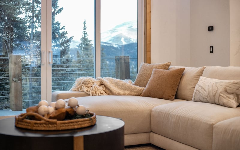 A cozy, neutral-toned sofa with textured pillows faces a large window showing a snowy mountain landscape.