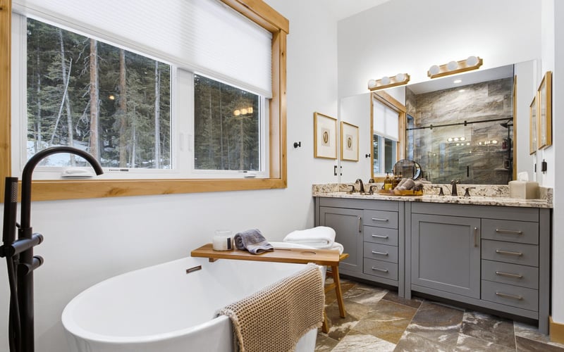 Modern bathroom with a freestanding white tub, grey vanity, stone flooring, and a large window overlooking a forest.