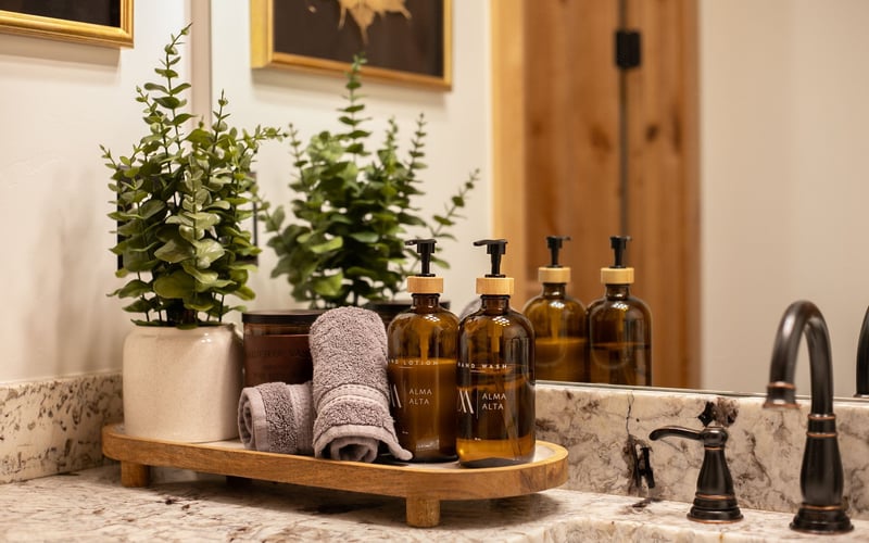 Amber soap bottles, rolled towels, and two potted plants rest on a wooden tray atop a marble bathroom vanity.