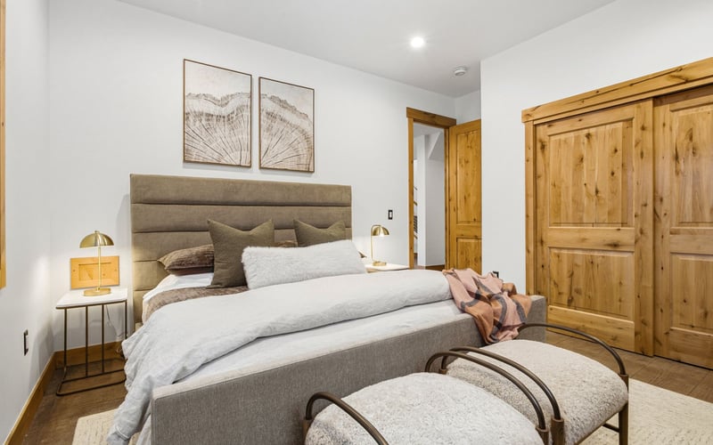 A modern bedroom features a gray upholstered bed, light bedding, two framed prints, wood sliding doors, and side benches.