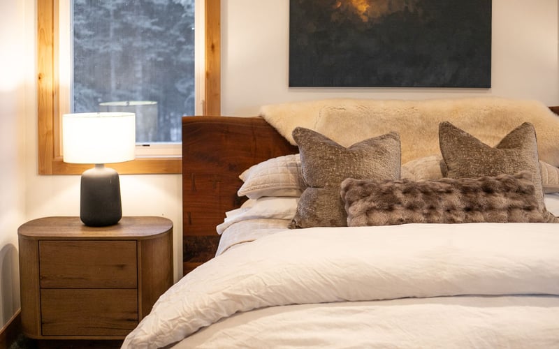 A cozy bedroom with a wooden nightstand, a lit table lamp, and a made bed featuring a white duvet and decorative pillows.