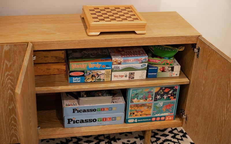 A wooden sideboard with an open cabinet, filled with board game boxes, topped with a small wooden chessboard.