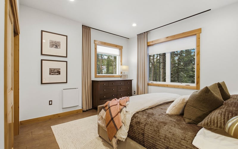 A cozy, well-lit bedroom featuring a bed with brown bedding, wooden accents, two framed prints, and a windowed dresser.
