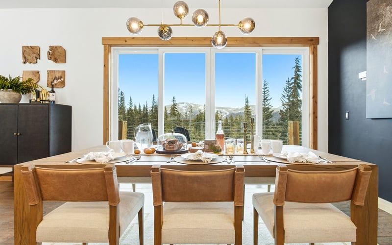 A dining room featuring a wooden table, three upholstered chairs, a modern chandelier, and a large window facing mountains.