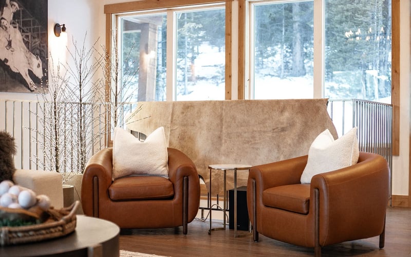 Two brown leather armchairs with white pillows face a window, with a small table between them in a living room.