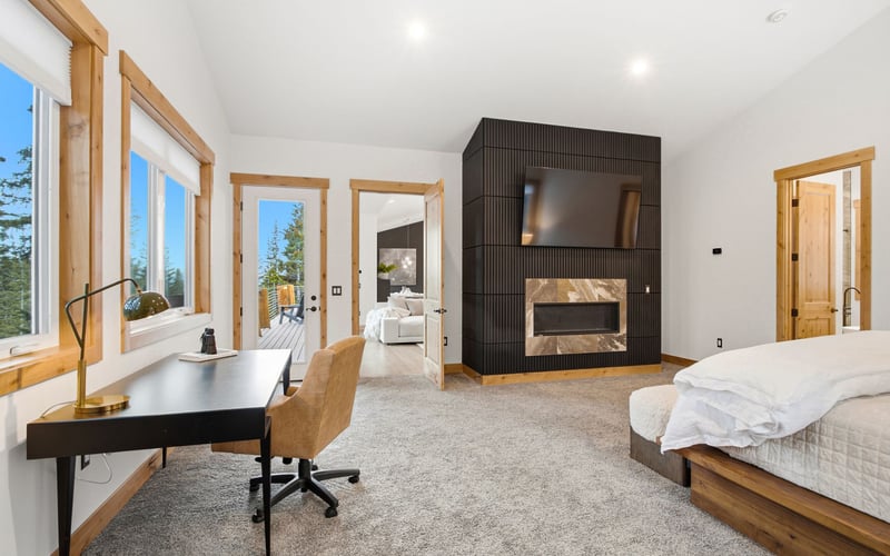 A modern bedroom with a desk, office chair, large fireplace with a mounted television, and a bed with white bedding.
