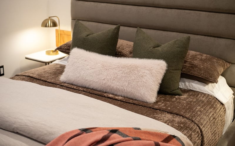 A bed with a tufted headboard, two dark olive pillows, a fuzzy cream lumbar pillow, and a patterned peach-toned blanket.