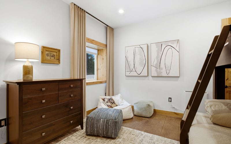 A bedroom with a wooden dresser, a window with beige curtains, a patterned ottoman, a beanbag chair, and bunk bed stairs.