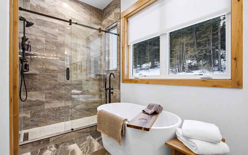A bright modern bathroom featuring a white soaking tub next to a large window and a stone-tiled walk-in glass shower.