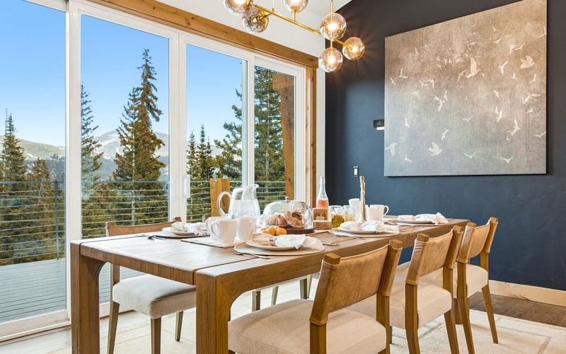 A dining room with a wooden table and chairs, set against a dark blue wall with art, overlooking a mountain forest.