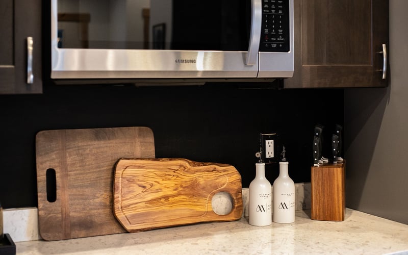 Kitchen counter with two wooden cutting boards, two white oil bottles, and a wooden knife block under a microwave.