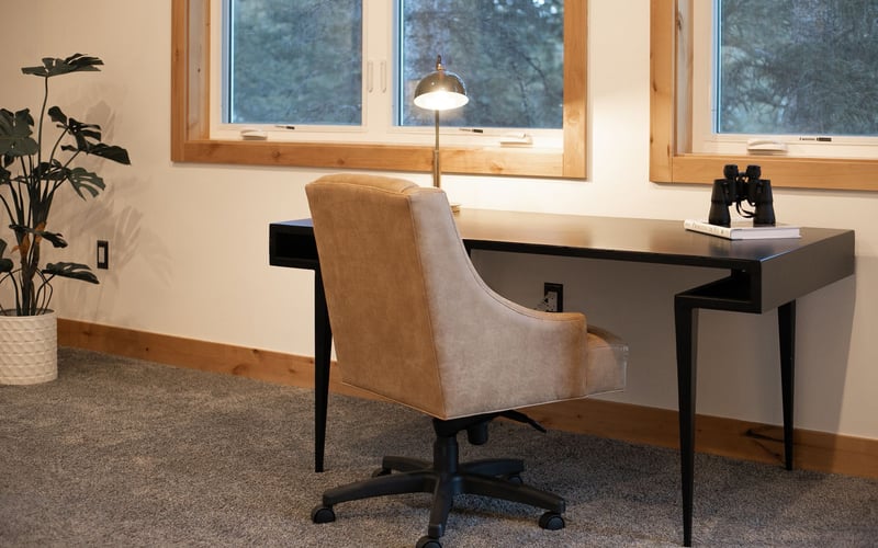 A modern workspace with a tan upholstered chair, a black desk with a lamp and binoculars, against a white wall.