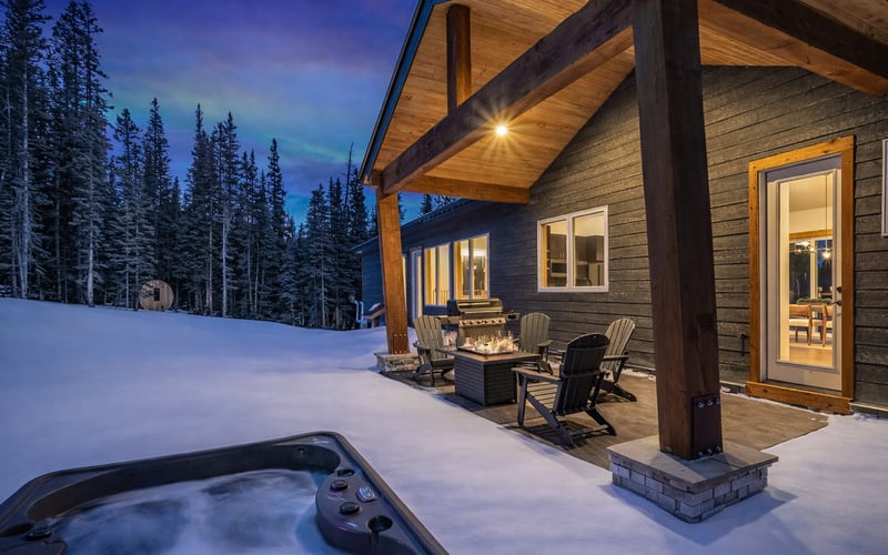A snowy patio at dusk with a hot tub, fire pit, and outdoor seating outside a modern cabin near a pine forest.