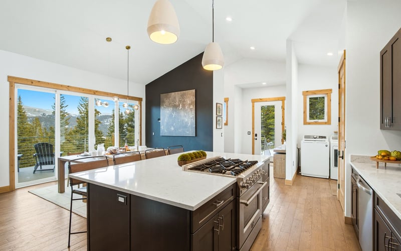 A modern kitchen with a large white island, dark cabinets, and a dining area overlooking a snowy mountain landscape.