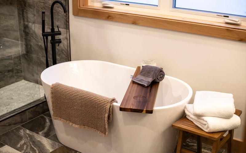 A white freestanding bathtub sits in a bathroom with dark tiled flooring, a wooden tray, and a stool with white towels.