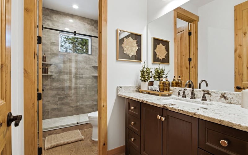 A modern bathroom featuring a dark wood vanity with a stone countertop, framed wall art, and an adjacent walk-in shower.