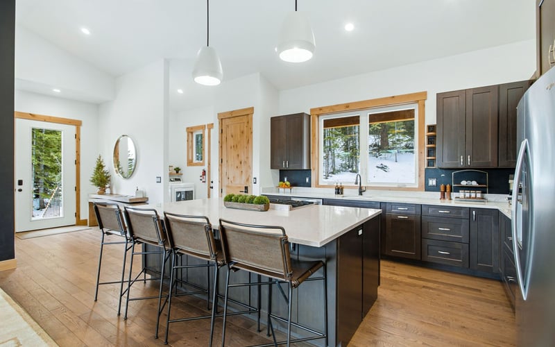 A modern kitchen with a large island, three stools, dark brown cabinets, white countertops, and wood floors.