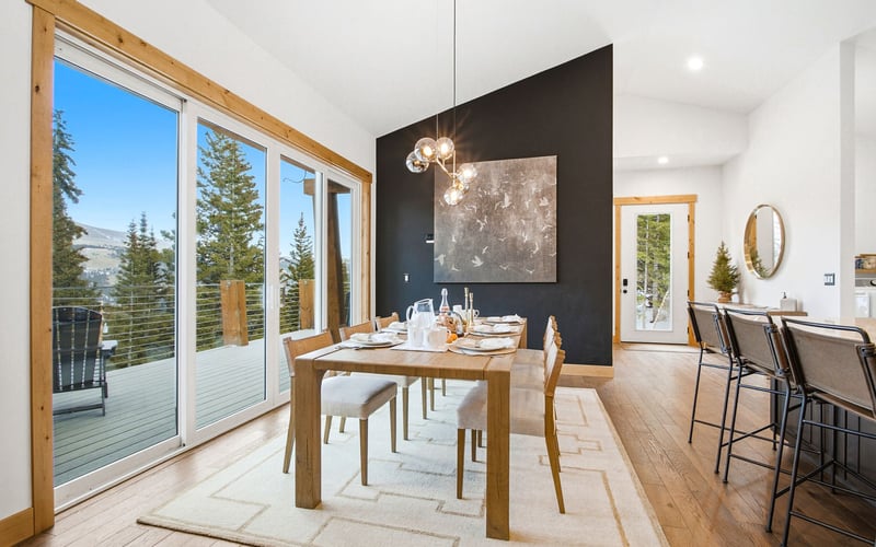 A modern dining room with a wooden table, grey chairs, pendant lights, and large sliding glass doors overlooking trees.