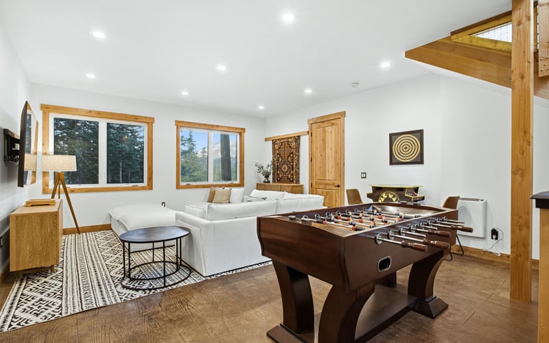 A bright, modern basement lounge with a foosball table, white sofa, patterned rug, and wooden accents.