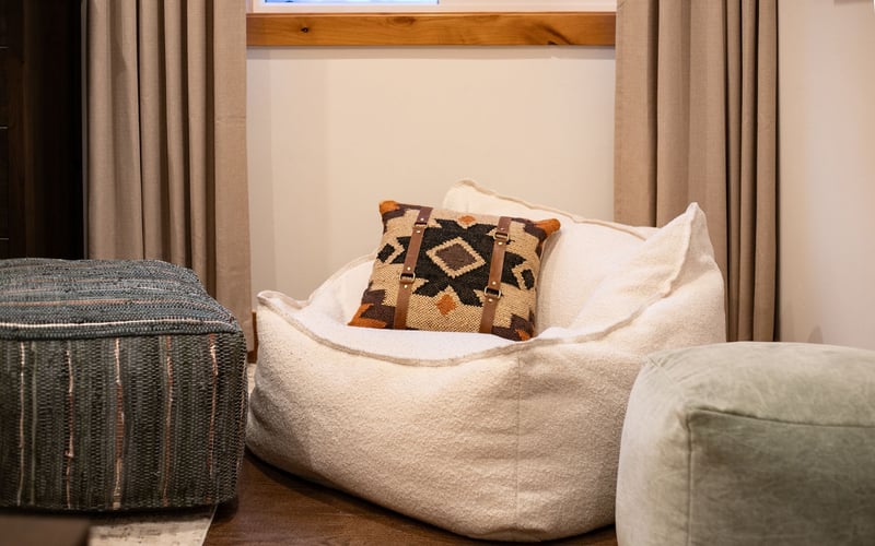 A fluffy white beanbag chair with a patterned throw pillow sits between two fabric ottomans in front of beige curtains.