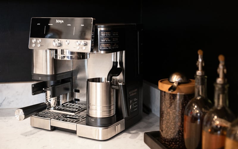 A stainless steel Ninja espresso machine on a marble countertop next to a container of coffee beans and glass dispensers.