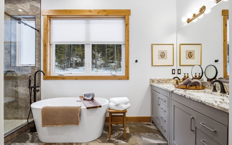 Modern bathroom with a freestanding tub, a walk-in stone shower, a gray double vanity, and a large window facing trees.