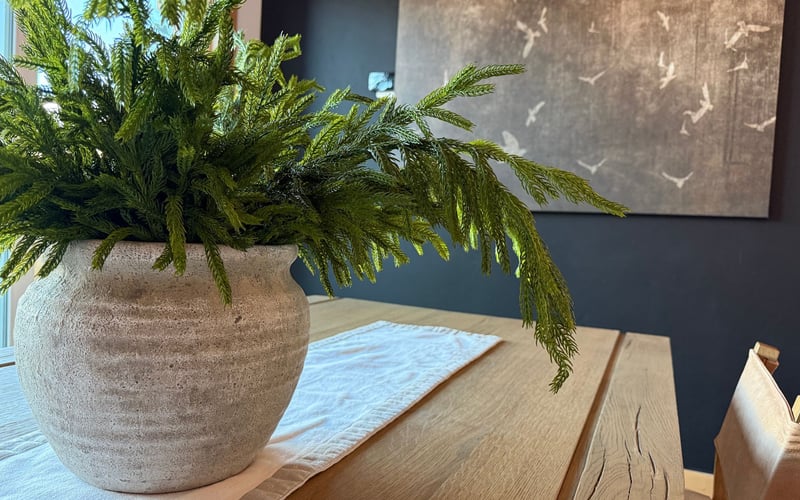 A textured, light-colored stone planter with a lush green plant sits on a light wood table with a white runner.