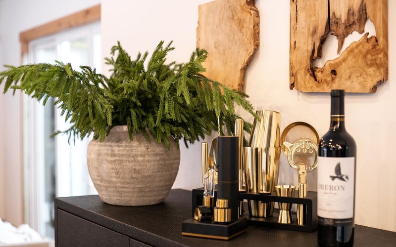A potted fern, gold bar tools, and a bottle of Heron wine sit on a black table against a wall with wood wall art.