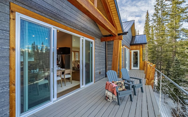 A modern wooden balcony with two blue chairs and sliding glass doors overlooking a snowy, tree-lined landscape.