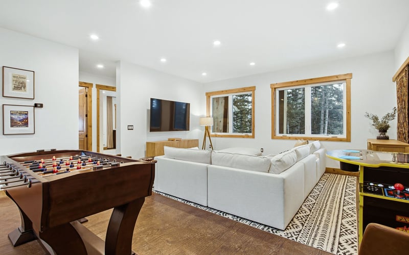 A modern living room with a white sectional sofa, a foosball table in the foreground, and large windows.