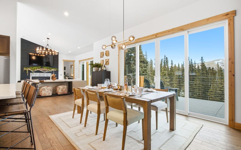 A modern dining room with a wooden table, six chairs, and a chandelier, next to sliding glass doors overlooking a forest.