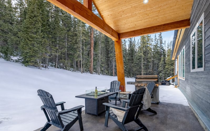 A covered patio featuring two dark Adirondack chairs, a fire pit table, and a grill, overlooking a snowy, pine-forested hill.