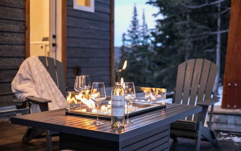 Two Adirondack chairs sit on a deck flanking a fire table with a wine bottle and two glasses, overlooking a snowy forest.