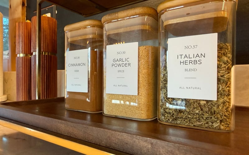 Three square glass spice jars with wooden lids labeled Cinnamon, Garlic Powder, and Italian Herbs on a wooden tray.